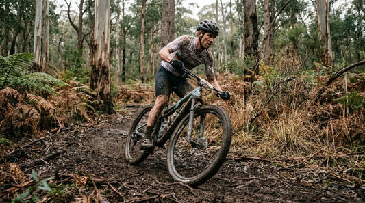 A mud-splattered mountain biker pinning through technical roots on a dark, loamy Australian trail during a winter race.