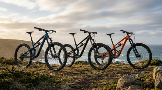 Three premium 2026 carbon mountain bikes lined up on a Tasmanian coastal cliff trailhead at golden hour overlooking the Southern Ocean