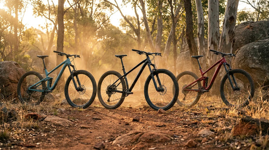 Three modern mountain bikes lined up at golden hour on red dirt singletrack in the Australian bush — full-suspension trail bike, hardtail, and dual-sus enduro options under $2500