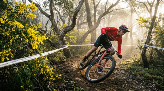 A mountain biker racing through a fast, loamy corner on an Australian bush trail with yellow wattle and morning mist.