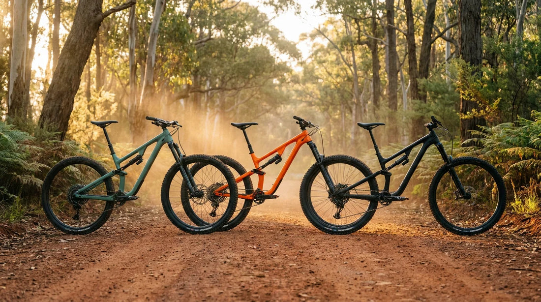 Three 2026 full-suspension trail mountain bikes lined up on a red dirt trailhead in the Australian eucalyptus bush at golden hour