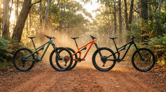 Three 2026 full-suspension trail mountain bikes lined up on a red dirt trailhead in the Australian eucalyptus bush at golden hour
